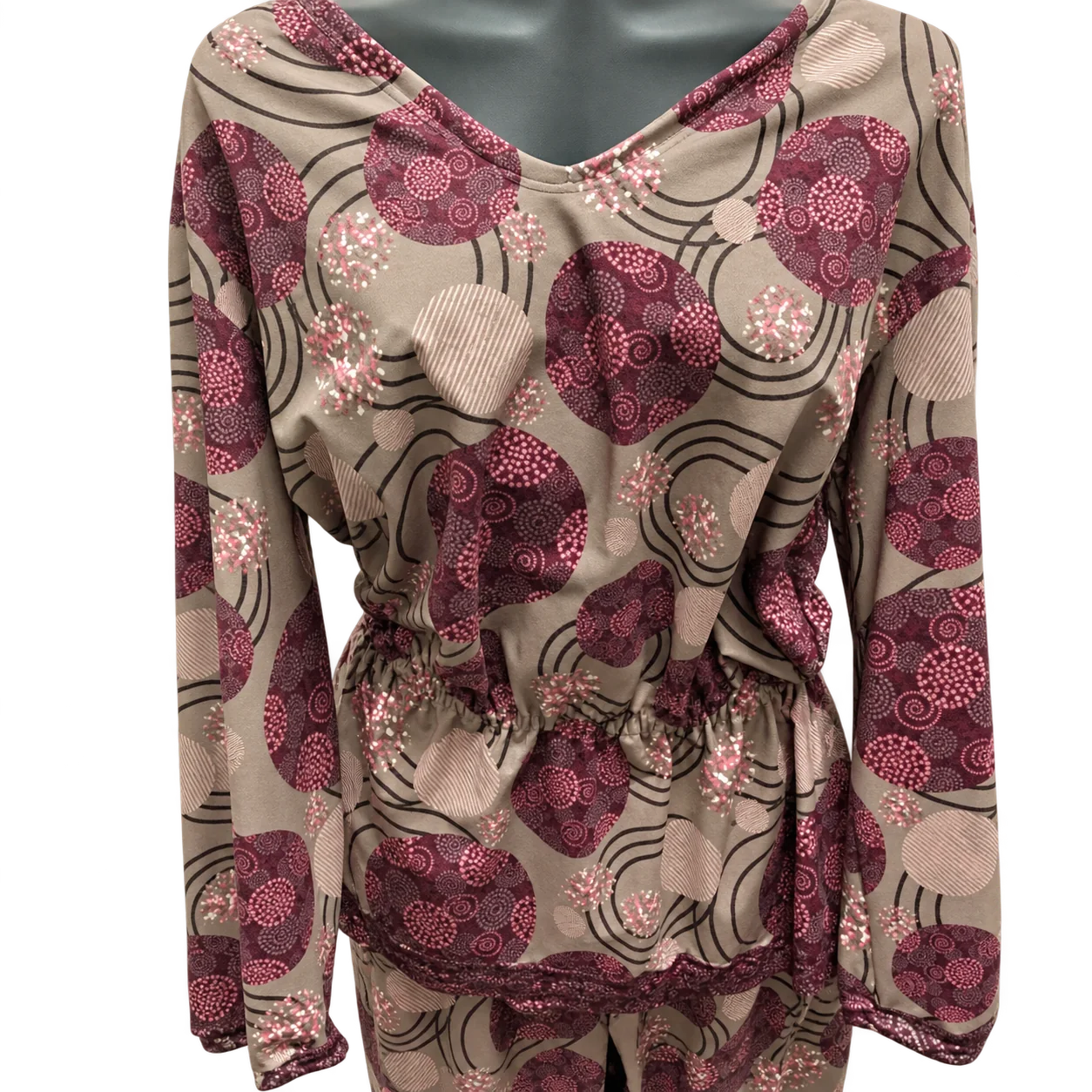 Une blouse à manches longues et col en V, ornée de grands motifs circulaires bordeaux et roses sur fond taupe, présentée sur un mannequin devant un mur clair.