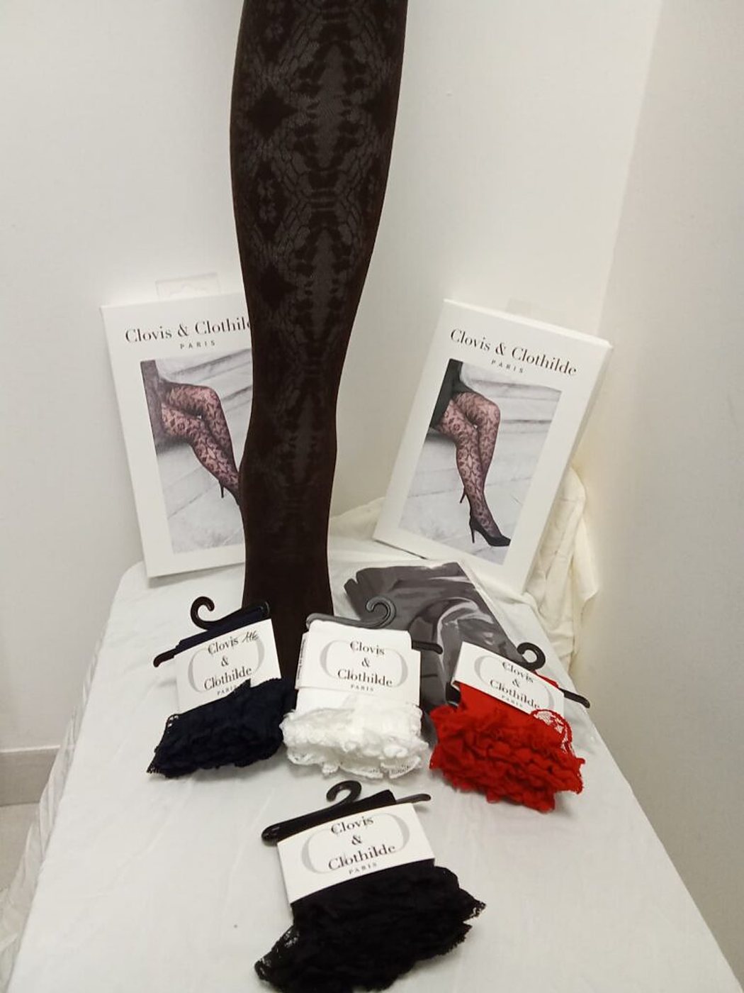 Un collant brun à motifs est présenté debout derrière deux boîtes de collants et quatre paires de socquettes en dentelle bleu marine, blanche, rouge et noire de la marque Clovis & Clothilde, posées sur une table drapée de blanc.
