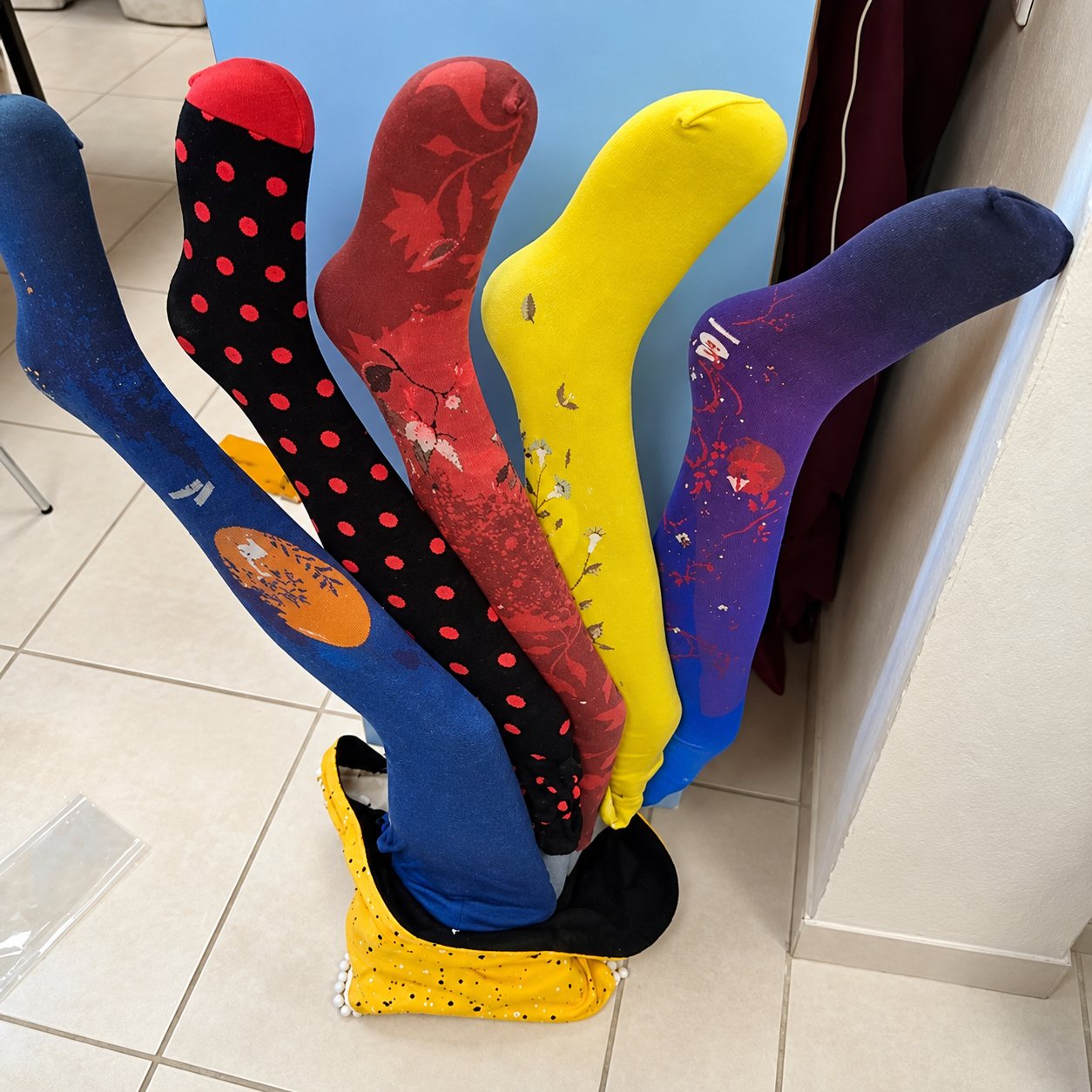 Cinq collants colorés, bleu, noir à pois rouges, bordeaux fleuri, jaune et violet, présentés sur des jambes de mannequin dressées sur un carrelage clair.