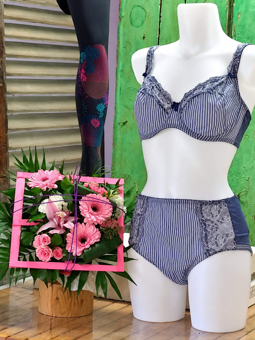Un mannequin blanc porte un ensemble de lingerie rayé bleu et blanc avec dentelle, à côté d'un bouquet de gerberas et de roses roses dans un cadre rose.