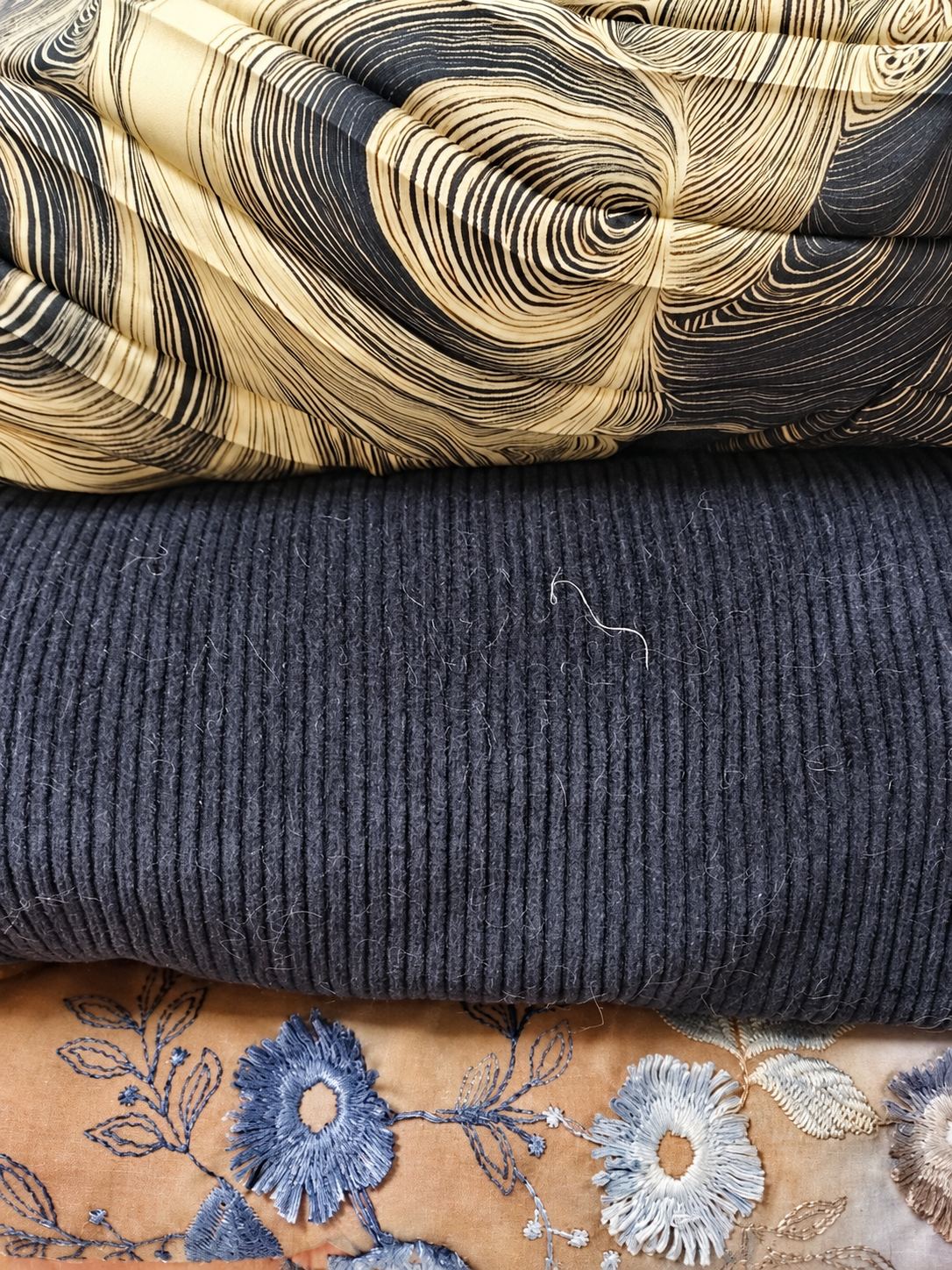 Trois tissus empilés : un tissu à motif ondulé noir et crème, un velours côtelé bleu marine et un tissu brodé de fleurs bleues sur fond beige.