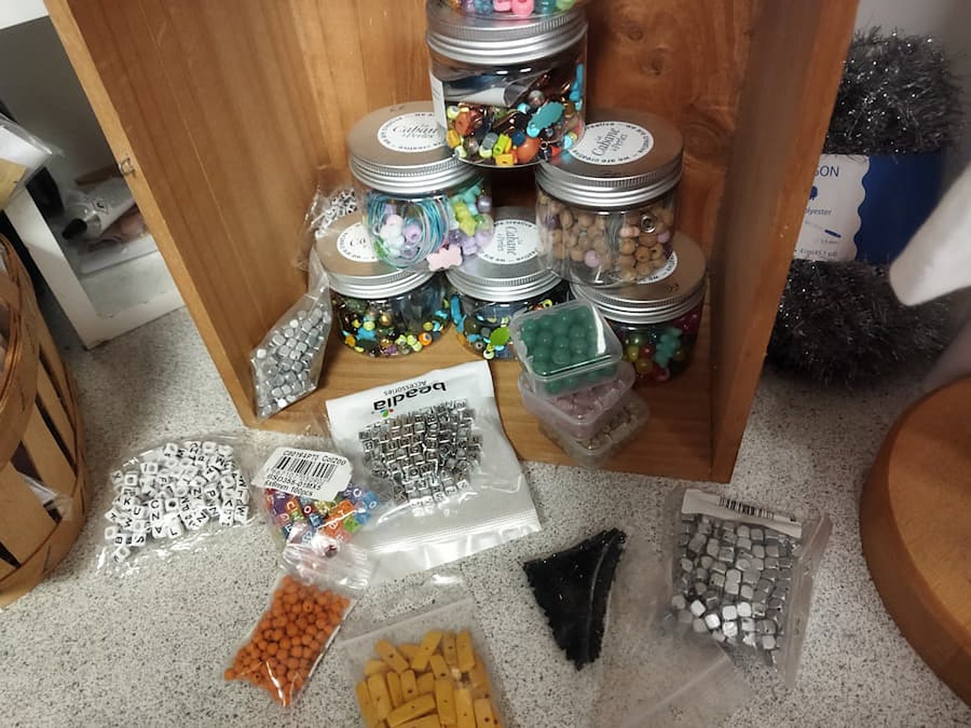 Des pots de perles multicolores sont empilés dans une caisse en bois, entourés de sachets de perles alphabet, argentées, orange et jaunes posés sur un plan de travail moucheté.