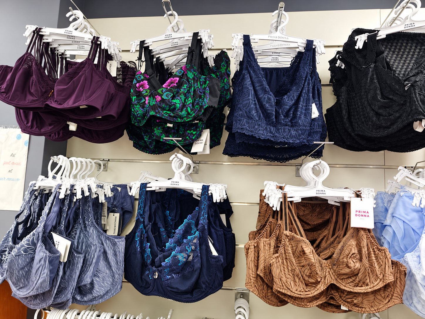 Des soutiens-gorge de différentes couleurs et motifs, sur cintres blancs Prima Donna, sont accrochés à un mur de présentation dans une boutique de lingerie.