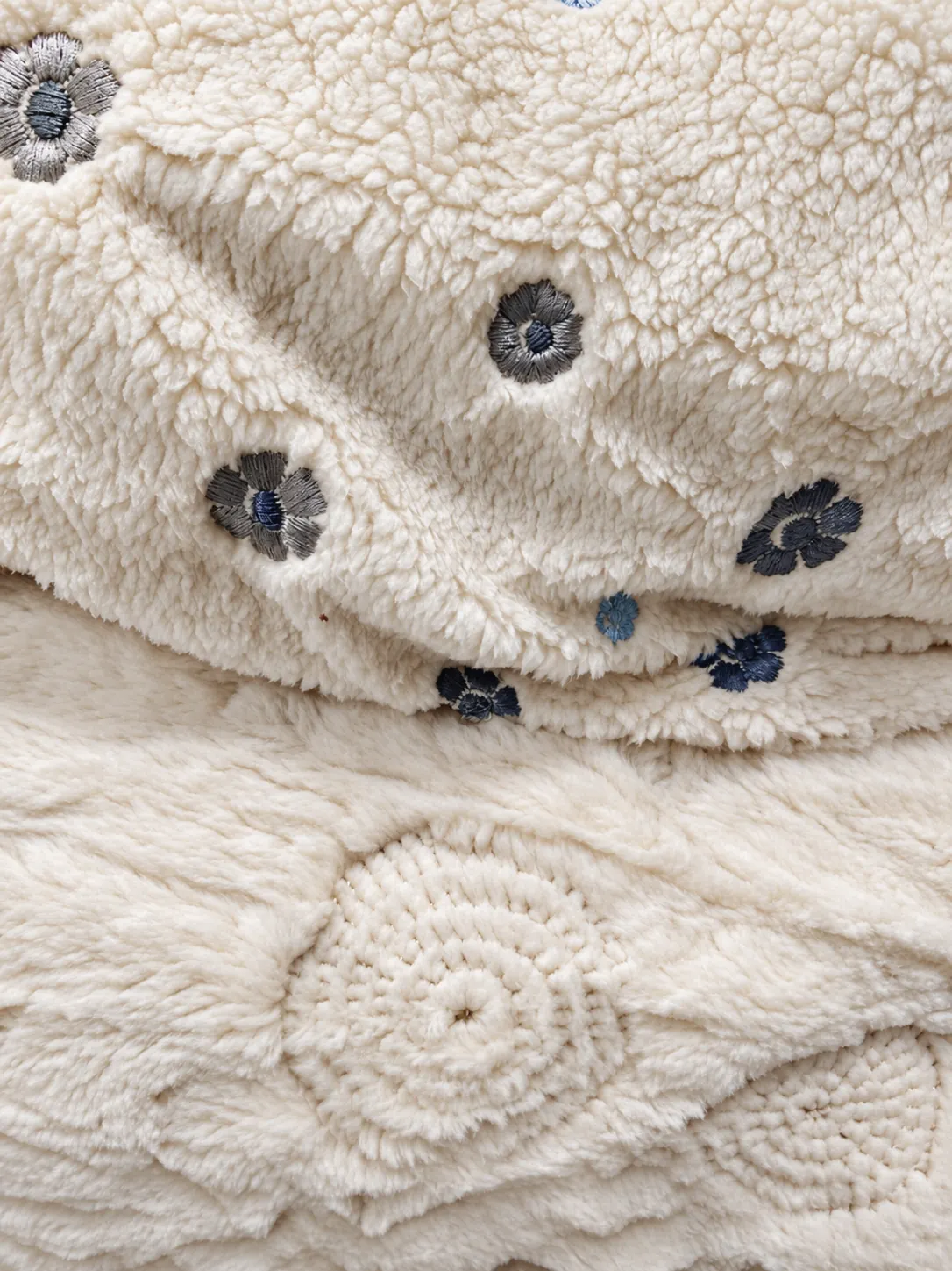 Gros plan sur un tissu sherpa écru et douillet orné de petites fleurs brodées grises et bleues, avec un tissu en relief à motifs circulaires en dessous.