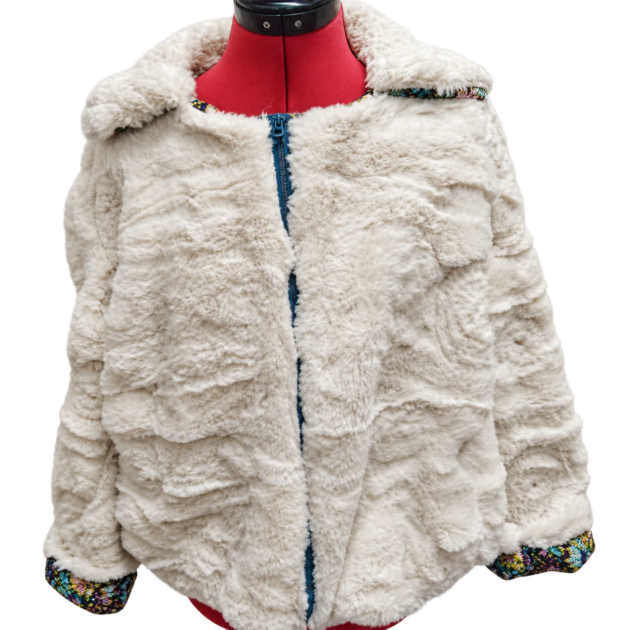 Une veste courte en fausse fourrure blanche à doublure colorée est présentée sur un mannequin de couture rouge devant un mur clair.